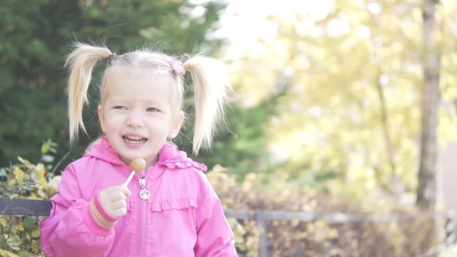 Little girl sucks chupa chups outside in the park. Close-up funny baby with two tails. A girl in a pink jacket holding a candy in her hands, takes it in her mouth and licks