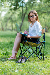 Business womanl with a laptop sitting in the Park on a folding chair and writing in a notebook