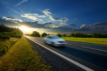 Motion blurred white passenger car driving on asphalt road in rural landscape at sunset