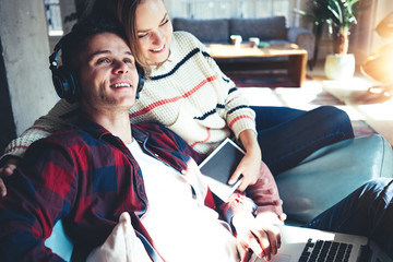 Happy couple at home. Husband and wife sitting on the couch together and watching TV series on laptop, listening music and reading books. Bright loft apartment