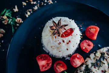 Basmati rice close up. Anise star, chili pepper, cherry tomatoes, chiken meat. Dark background, black plate