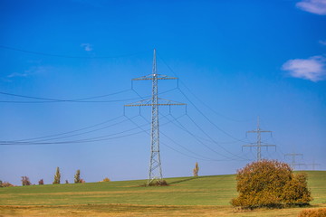 Bright blue sky, white clouds, horizon with trees, high voltage electricity pylons standing on field, green grass, yellow and brown foreground, bush
