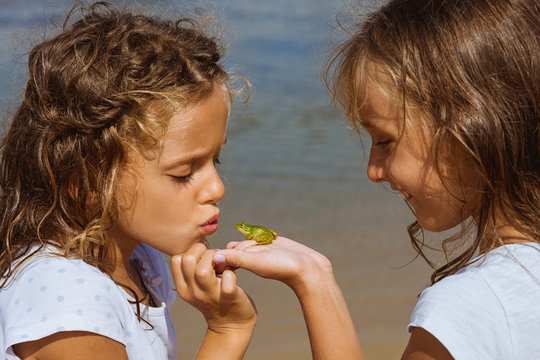 Photography of two beautiful twin girls by the sea, with a frog in their hands. One girl is holding the green baby frog and smiles. The other girl is kissing the animal.