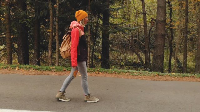 A young slim girl in a yellow hat and a vintage backpack walks along an asphalt road in the autumn yellow coniferous forest. Side view