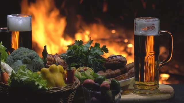 Two Beers, vegetables and grilled sausages on fire background, dolly shot