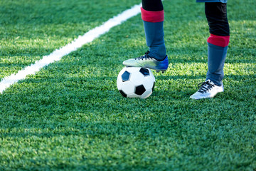 Obraz premium classical black and white football ball on the green grass of the field and leg in sports shoes on it. Soccer game, training, hobby concept. with copy space 