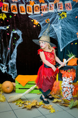 A child, a little girl in the shape of a witch on a broomstick, poses against the backdrop of scenery of cobwebs, pumpkins and autumn leaves on a Halloween holiday.