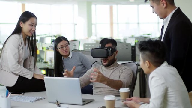 Medium shot of Asian office worker using virtual reality headset when sitting at desk surrounded by group of colleagues