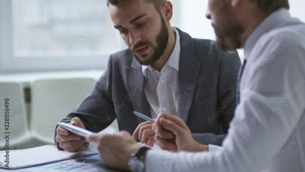 Poster tilt up shot of two men in formal clothes talking to each other when sitting at office desk and usin