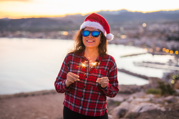 Holiday, Christmas and people concept - Young happy woman in Santa hat over beautiful landscape with fireworks and sparklers