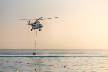 A helicopter with a red basket descends over the sea to scoop water against the background of the dawn orange sky and the silhouette of the city in the distance