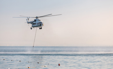 A helicopter with a red basket descends over the sea to scoop water against the background of the dawn orange sky and the silhouette of the city in the distance