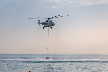 A helicopter with a red basket descends over the sea to scoop water against the background of the dawn orange sky and the silhouette of the city in the distance