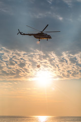 A fire helicopter collects water into the sea and flies toward the mountains to extinguish a forest fire on the background of a beautiful dawn sky. Rescue operation. Forest Fire Prevention