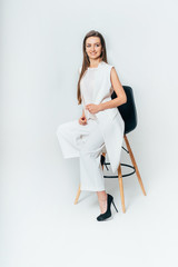 Young business woman sits on a chair in the studio on a white background. A woman in a white suit and black shoes looks to the side. Close-up