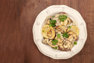 A photo of scallopine di vitello, veal scallopini, with a mushroom sauce, lemons, and parsley, shot from the top on a dark rustic background with copy space
