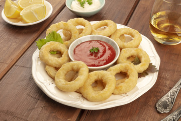 A closeup photo of squid rings with a tomato sauce and Mayonnaise, lemon wedges, and white wine, on a dark rustic wooden background