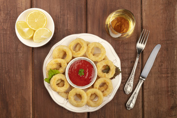 An overhead photo of squid rings with a red sauce, lemon wedges, white wine, and copy space