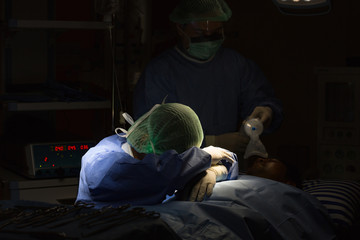 Asian surgeon resting head on arms beside patient during surgery, expressing emotional exhaustion...