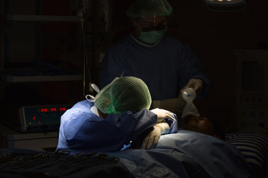 Asian surgeon resting head on arms beside patient during surgery, expressing emotional exhaustion under low lighting in operating room. - Powered by Adobe