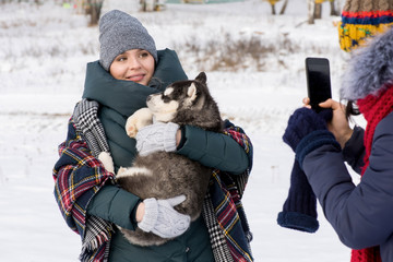 Waist up portrait of modern young woman holding cute Husky puppy and looking at smartphone camera with friend taking photo of her outdoors in winter