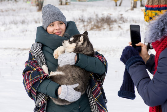 Waist up portrait of modern young woman holding cute Husky puppy and looking at smartphone camera with friend taking photo of her outdoors in winter