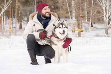 Full length portrait of modern Asian man posing with gorgeous Husky dog sitting down outdoors in winter landscape and smiling happily, copy space