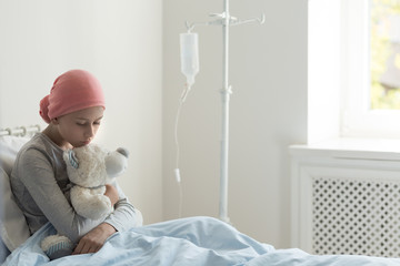 Sad ill teenager girl sitting alone in hospice bed, holding teddy bear, waiting for her doctor