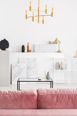 Vertical view of backside of powder pink sofa in white modern living room interior with coffee table, golden chandelier and maps in white frames, real photo