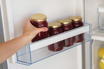 Obraz na płótnie Canvas Glass Jars with Raspberry Jam on shelf in fridge. Female hand with a jar of red homemade jam. Fermented healthy foods stand in refrigerator.
