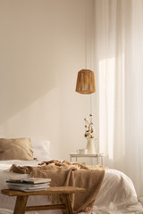 Wooden stool in front of bed with linen blanket in natural bedroom interior with lamp. Real photo