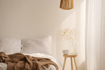 Plant on wooden stool next to bed with brown blanket in white simple bedroom interior. Real photo
