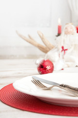 Christmas place setting with white dishware, cutlery, silverware and red decorations on wooden board. Christmas candlestick with red candles as decor.