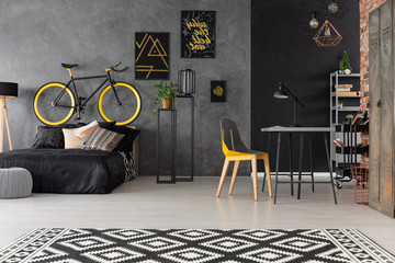 Patterned carpet and chair at desk in grey room interior with posters and bike above bed. Real photo