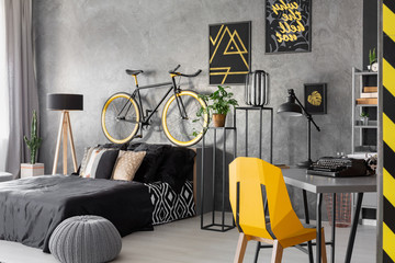 Posters and bike above black bed in grey teenager's room interior with pouf and yellow chair. Real photo