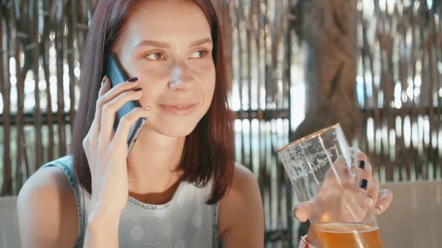 Young woman drinking beer and communicates by mobile phone in a street bar.