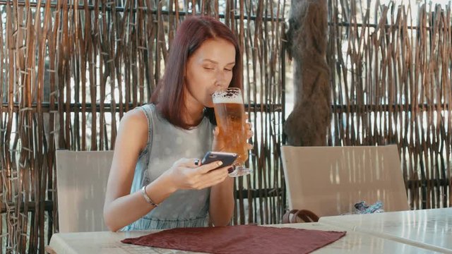 Young woman drinking beer and communicates by mobile phone in a street bar.