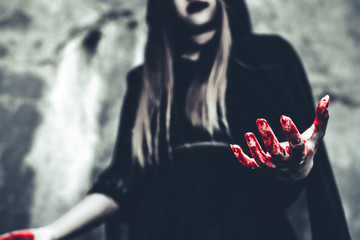 Witch showing bloody hand. Female demon angel in black clothes and hood on grunge wall background. Halloween day and Mystery concept. Fantasy of magic theme. Afterlife and Death concept. - obrazy, fototapety, plakaty