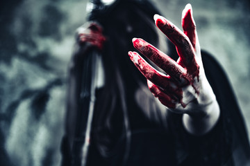 Witch showing bloody hand. Female demon angel in black clothes and hood on grunge wall background....