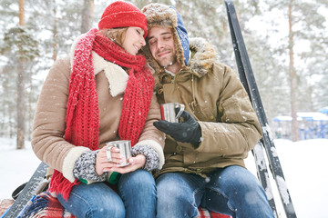 Obraz premium Portrait of loving young couple cuddling tenderly and enjoying hot cocoa during date in beautiful winter forest, copy space