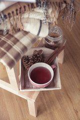 Plaids. A cup of hot tea. Autumn cozy interior. On a wooden chair is a stack of warm blankets. Candles, leaves, cones, basket, cinnamon. Book and glasses. Autumn. Winter.