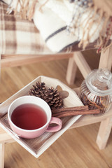Plaids. A cup of hot tea. Autumn cozy interior. On a wooden chair is a stack of warm blankets. Candles, leaves, cones, basket, cinnamon. Book and glasses. Autumn. Winter.