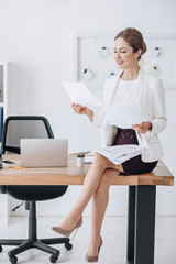 attractive professional business woman doing paperwork while sitting on table with laptop