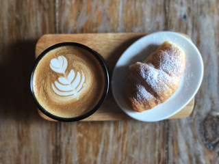 Delicious Breakfast;  Heart love Latte art coffee in Black cup and Croissant topped with icing sugar in white dish on the wooden try and vintage wooden table.