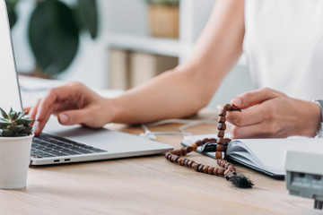 cropped view of businesswoman using laptop and holding rosary beads at workplace