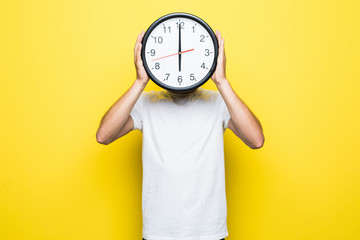 Young man holding big clock covering his face on yellow background - obrazy, fototapety, plakaty