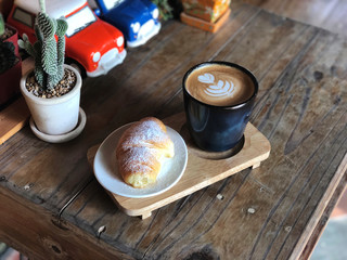 Delicious Breakfast;  Heart love Latte art coffee in Black cup and Croissant topped with icing sugar in white dish on the wooden try and vintage wooden table.