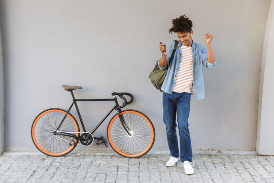 Happy young african guy walking outdoors with bicycle listening music chatting by mobile phone.