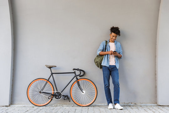 Happy young african guy walking outdoors with bicycle chatting by mobile phone.