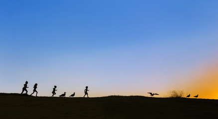 Obraz na płótnie Canvas Silhouettes of four running kids and seagulls on the horizon on sunset. Sky is blue, sun sets in the right corner. Lots of empty space, copy space, for design, meme, background, wallpaper, poster, ad.